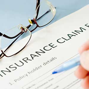 Close up of an insurance claim form with reading glasses on the top left and a person holding a blue pen on the right